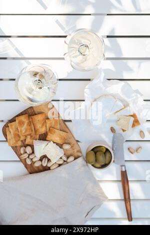 Deux verres de vin blanc et une assiette en bois avec du fromage et des noix sur une table blanche à l'extérieur. Banque D'Images