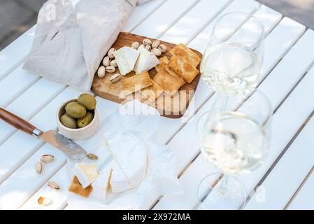 Deux verres de vin blanc et une assiette en bois avec du fromage et des noix sur une table blanche à l'extérieur. Banque D'Images