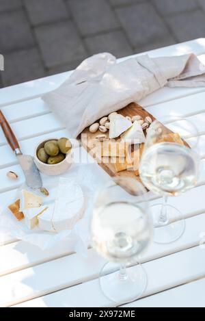 Deux verres de vin blanc et une assiette en bois avec du fromage et des noix sur une table blanche à l'extérieur. Banque D'Images