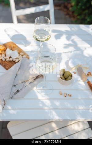 Deux verres de vin blanc et une assiette en bois avec du fromage et des noix sur une table blanche à l'extérieur. Banque D'Images