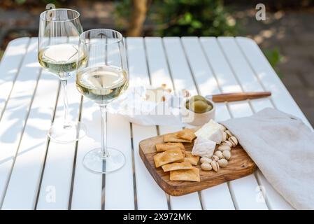 Deux verres de vin blanc et une assiette en bois avec du fromage et des noix sur une table blanche à l'extérieur. Banque D'Images