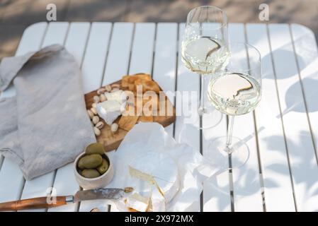 Deux verres de vin blanc et une assiette en bois avec du fromage et des noix sur une table blanche à l'extérieur. Banque D'Images