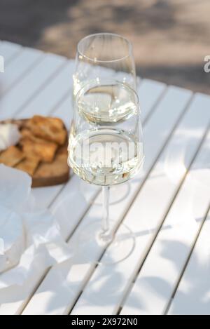 Deux verres de vin blanc et une assiette en bois avec du fromage et des noix sur une table blanche à l'extérieur. Banque D'Images