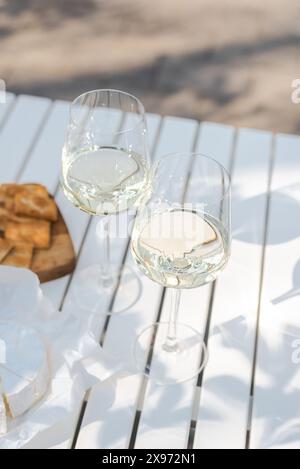 Deux verres de vin blanc et une assiette en bois avec du fromage et des noix sur une table blanche à l'extérieur. Banque D'Images