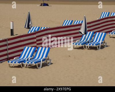 Plage où transats rayés bleus et blancs, parasols et cloisons rouges sont disposés en rangées, chaises longues colorées en bord de mer avec ciel bleu Banque D'Images