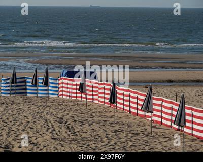 Large plage de sable avec des parasols bleus et blancs rayés et noirs dispersés et la mer avec des vagues en arrière-plan, chaises de plage colorées par Banque D'Images