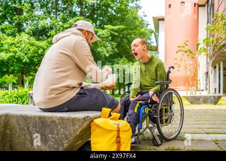 Étudiant universitaire adulte caucasien masculin nourrir un ami atteint de paralysie cérébrale assis sur un fauteuil roulant dans le campus Banque D'Images