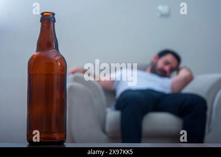 Bouteille d'alcool en gros plan tandis que l'homme dormant sur le canapé en arrière-plan fuyant de l'alcool Banque D'Images