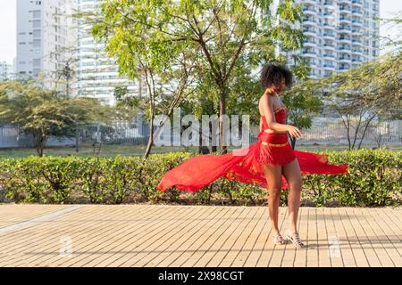 Une femme dans une robe rouge vibrante danse gracieusement dans un parc urbain avec de la verdure et des immeubles de grande hauteur en arrière-plan, mettant en valeur un moment animé. Banque D'Images