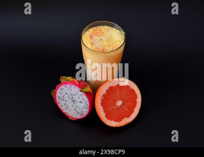 Un grand verre à facettes de smoothie aux fruits sur un fond noir, à côté des moitiés de pitaya mûr et de pamplemousse. Gros plan. Banque D'Images