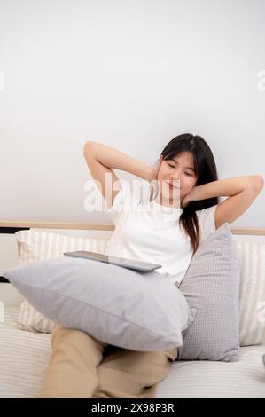 Une jeune femme asiatique attrayante, portant des vêtements confortables, étire ses mains après avoir utilisé sa tablette numérique alors qu'elle était assise dans un lit confortable Banque D'Images