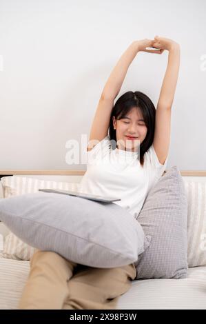 Une jeune femme asiatique attrayante, portant des vêtements confortables, étire ses mains après avoir utilisé sa tablette numérique alors qu'elle était assise dans un lit confortable Banque D'Images