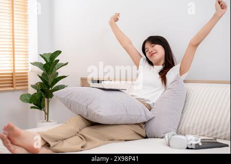 Une jeune femme asiatique attrayante, portant des vêtements confortables, étire ses mains après avoir utilisé sa tablette numérique alors qu'elle était assise dans un lit confortable Banque D'Images