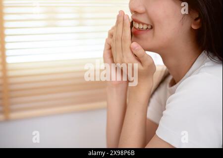 Une image recadrée d'une jeune femme asiatique joyeuse couvrant sa bouche tout en parlant au téléphone avec son amie à l'intérieur, appréciant parler ou commérer o Banque D'Images