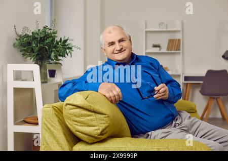 Portrait d'un homme âgé assis sur un canapé dans le salon à la maison et tenant des lunettes dans les mains. Banque D'Images
