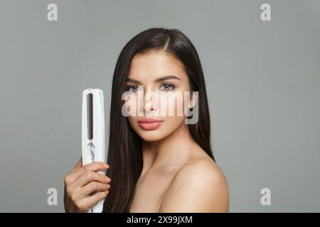 Élégante jeune femme brune saine tenant le fer à cheveux et lissant ses longs cheveux lisses brillants sur fond blanc. Soins capillaires, coiffure, cheveux Banque D'Images