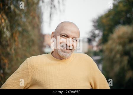 Homme âgé dans un pull jaune, souriant doucement, profitant de l'environnement extérieur serein dans un parc urbain luxuriant Banque D'Images