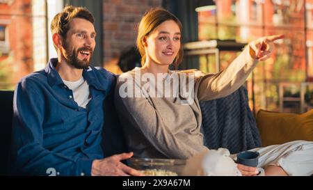 Portrait de jeune couple passant du temps à la maison, assis sur un canapé, l'homme embrassant sa petite amie tout en regardant l'émission de télévision passionnante dans l'appartement. Homme et femme streaming film et soyez surpris. Banque D'Images