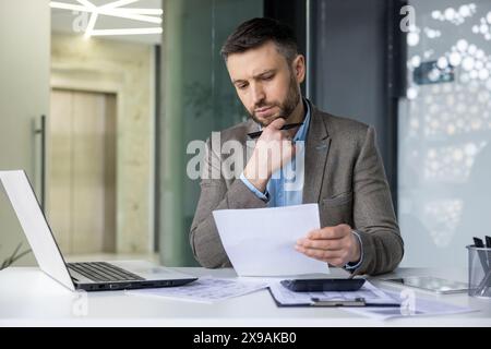 Un homme d'affaires profondément concentré sur l'analyse de documents à son bureau dans un bureau moderne. Il semble réfléchi et contemplatif tout en tenant un stylo. Banque D'Images