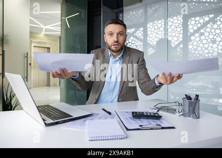 Homme d'affaires confus tenant des documents dans le bureau tout en étant assis au bureau avec ordinateur portable, calculatrice et stylos Banque D'Images