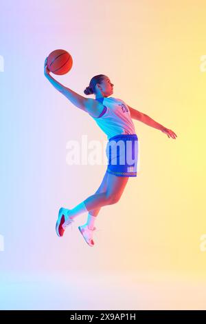 Portrait d'une joueuse professionnelle de basket-ball sautant avec la balle prête à marquer le but dans la lumière néon sur fond de studio dégradé. Banque D'Images