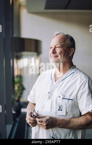 Médecin masculin senior attentionné dans les gommages tenant des lunettes tout en se tenant debout à l'hôpital Banque D'Images