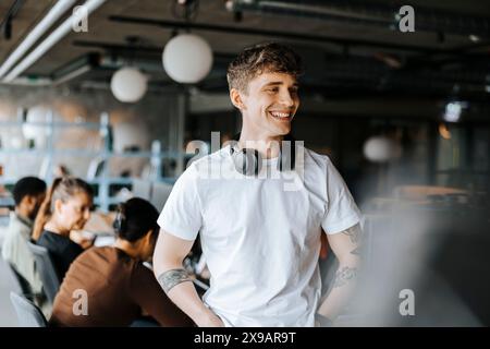 Jeune entrepreneur masculin souriant regardant loin tout en se tenant debout au bureau Banque D'Images