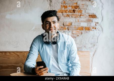 Portrait d'entrepreneur masculin souriant tenant un téléphone intelligent tout en étant assis contre un mur de briques au bureau Banque D'Images