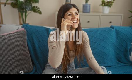 Belle jeune femme hispanique parlant au téléphone tout en étant assise sur un canapé couvert de bleu dans un salon confortable avec des plantes d'intérieur et pilule décorative Banque D'Images