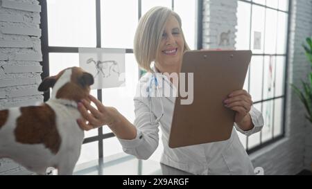 Une femme blonde vétérinaire d'âge moyen examine un petit chien dans une clinique vétérinaire d'intérieur, souriant tout en tenant une planche à pince. Banque D'Images