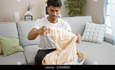 Beau jeune homme hispanique pliant la lessive sur un canapé dans un salon d'appartement confortable. Banque D'Images