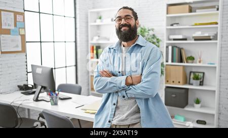 Homme hispanique barbu souriant dans une tenue décontractée se tient en toute confiance dans un bureau moderne avec les bras croisés. Banque D'Images