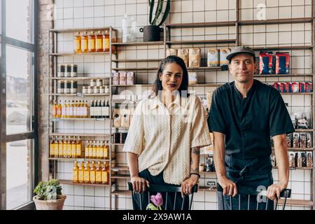 Portrait de propriétaire féminin debout avec un collègue masculin appuyé sur une chaise contre une étagère de marchandise au café Banque D'Images