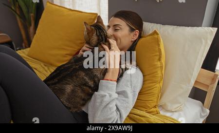 Une jeune femme hispanique se relaxant au lit à la maison avec un chaton, partageant un moment tendre dans une chambre confortable avec des oreillers jaunes et un éclairage doux. Banque D'Images