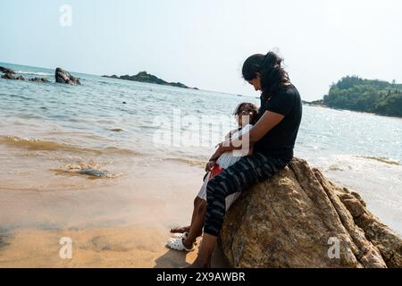 Un personnage solitaire est assis sur un éperon rocheux, perdu dans la pensée, tandis que de douces vagues embrassent le rivage sablonneux. Un moment de réflexion tranquille. Banque D'Images