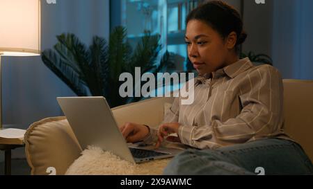 Focused afro-américaine femme ethnique fille 30s femme travaillant avec un ordinateur portable sur un canapé confortable utilisez un appareil informatique sur le canapé naviguant sur Internet social Banque D'Images