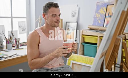 Bel homme hispanique tenant un café dans un studio d'art, entouré de peintures et d'un intérieur lumineux. Banque D'Images