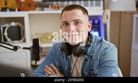 Beau jeune homme avec des écouteurs sourit en toute confiance dans une configuration de studio de musique, avec un microphone et des instruments en arrière-plan. Banque D'Images