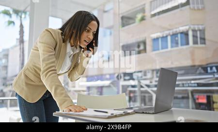 Une femme hispanique professionnelle en costume beige utilise un smartphone et un ordinateur portable dans un bureau moderne avec une vue sur le paysage urbain. Banque D'Images