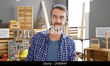 Un homme barbu d'âge moyen portant une chemise à carreaux pose dans un atelier avec des outils de travail du bois et des caisses en bois. Banque D'Images