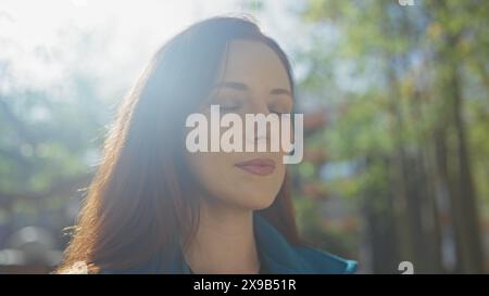 Une jeune femme sereine profitant d'un moment paisible dans un parc urbain ensoleillé, embrassant la tranquillité de la nature au milieu de la vie urbaine. Banque D'Images