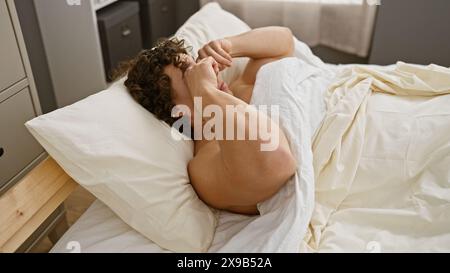 Un jeune homme hispanique aux cheveux bouclés est allongé dans le lit, paraissant stressé ou submergé dans une chambre minimaliste. Banque D'Images