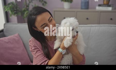 Une jeune femme hispanique souriante tenant un chien maltais bichon blanc dans un cadre confortable de salon Banque D'Images