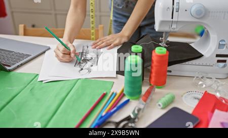 Une femme esquisse un dessin de robe sur papier au milieu de tissus colorés, de fils et d'une machine à coudre dans un atelier de tailleur. Banque D'Images