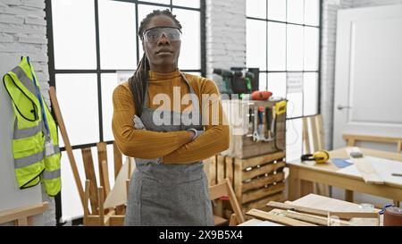 Femme confiante avec des tresses portant des lunettes de sécurité se croise les bras dans un atelier intérieur. Banque D'Images