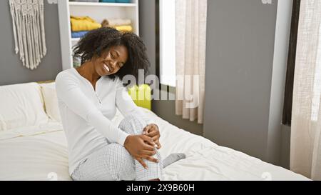 Femme afro-américaine souriant chaleureusement tout en étant assise sur le lit dans un intérieur de chambre moderne. Banque D'Images