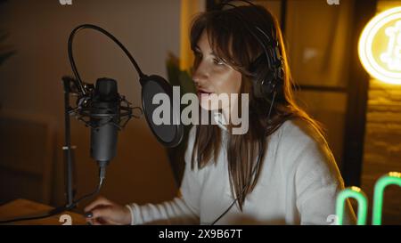 Une jeune femme portant des écouteurs parle dans un microphone dans un studio de radio intime, suggérant une émission en direct ou un scénario d'enregistrement de podcast Banque D'Images