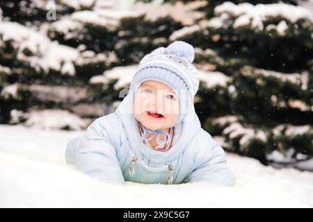 Un bébé dans une veste bleue et un bonnet en tricot profitant de la neige à l'extérieur avec une toile de fond d'arbres enneigés Banque D'Images