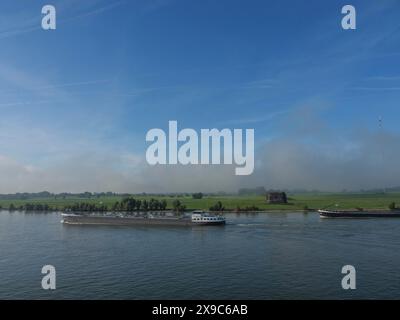 Bateaux naviguant le long d'une rivière, bordés par des prairies verdoyantes et un ciel brumeux avec un vieux bâtiment en arrière-plan, trafic maritime sur le Rhin à proximité Banque D'Images