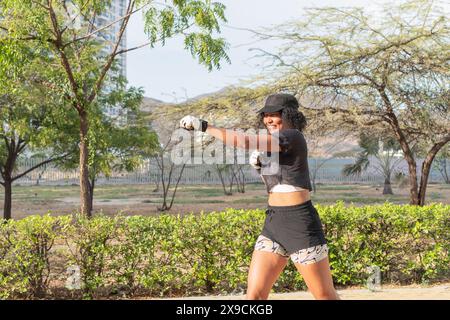 Femme pratiquant la boxe en plein air, portant une casquette et des vêtements de sport, souriant tout en frappant en avant dans un cadre de parc ensoleillé. Banque D'Images